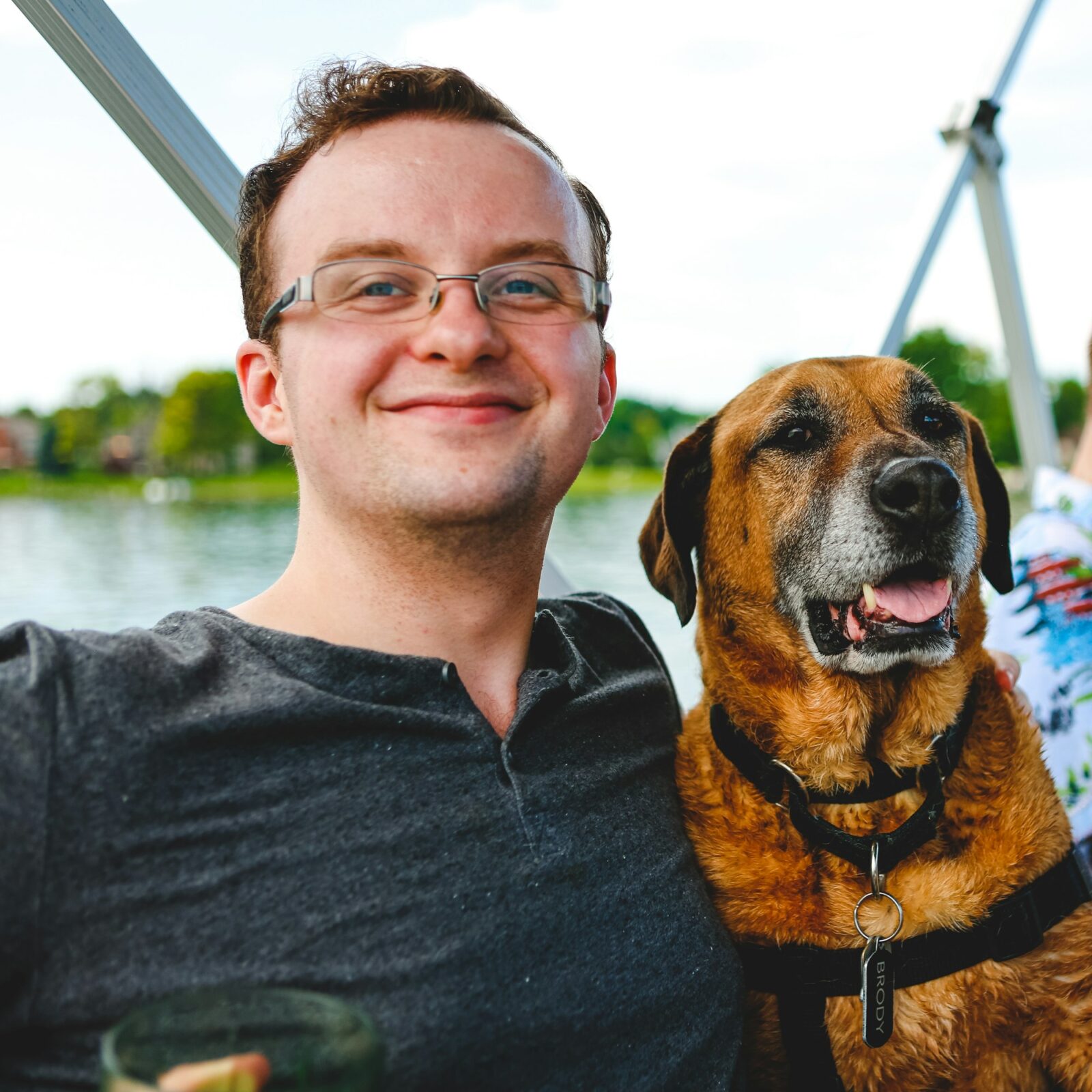 Man in glasses smiling at the camera and hugging a big dog with his tongue out, while on a boat